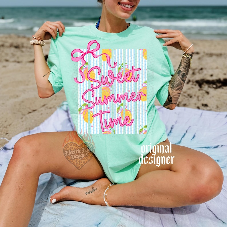 May include: A light green t-shirt with the words "Sweet Summer Time" in pink script. The design includes a pink bow and lemons on a blue and white striped background. The shirt is a relaxed fit.