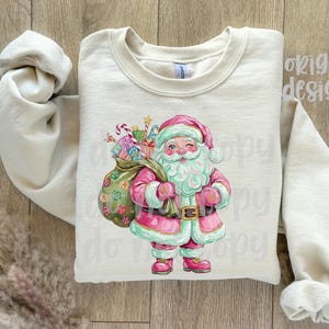 May include: Cream-coloured sweatshirt with a watercolour illustration of Santa Claus. Santa is in a pink suit with a matching hat and boots, holding a sack of gifts. The design includes candy canes and wrapped presents, perfect for the holiday season.