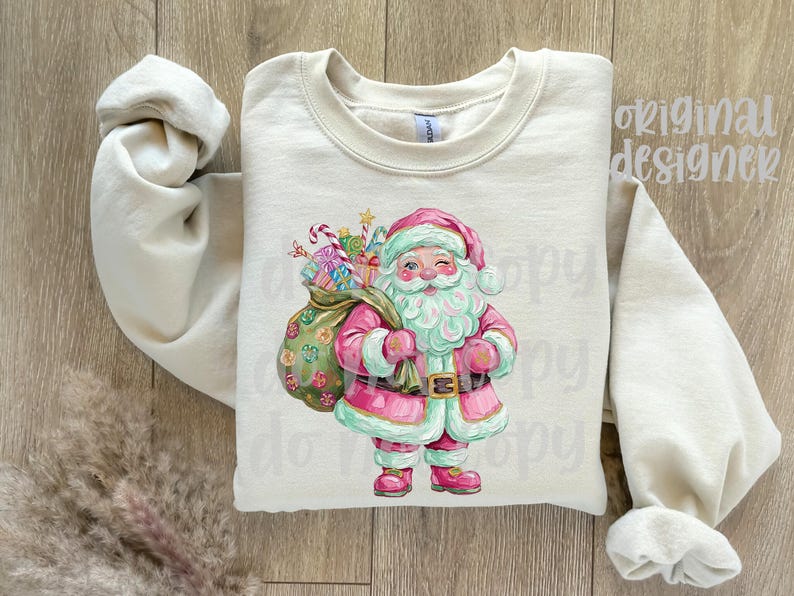 May include: Cream-coloured sweatshirt with a watercolour illustration of Santa Claus. Santa is in a pink suit with a matching hat and boots, holding a sack of gifts. The design includes candy canes and wrapped presents, perfect for the holiday season.