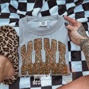 May include: A light gray sweatshirt with the word "MAMA" in a leopard print design. The sweatshirt is folded and resting on a black and white checkered blanket. A leopard print slipper is visible on the left.