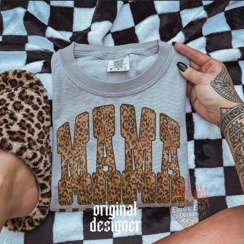 May include: A light gray sweatshirt with the word "MAMA" in a leopard print design. The sweatshirt is folded and resting on a black and white checkered blanket. A leopard print slipper is visible on the left.