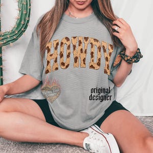 May include: A gray t-shirt with the word "HOWDY" in a brown and tan animal print design. The shirt also features a heart-shaped graphic with the text "Electric Edge Designs" and the words "original designer".
