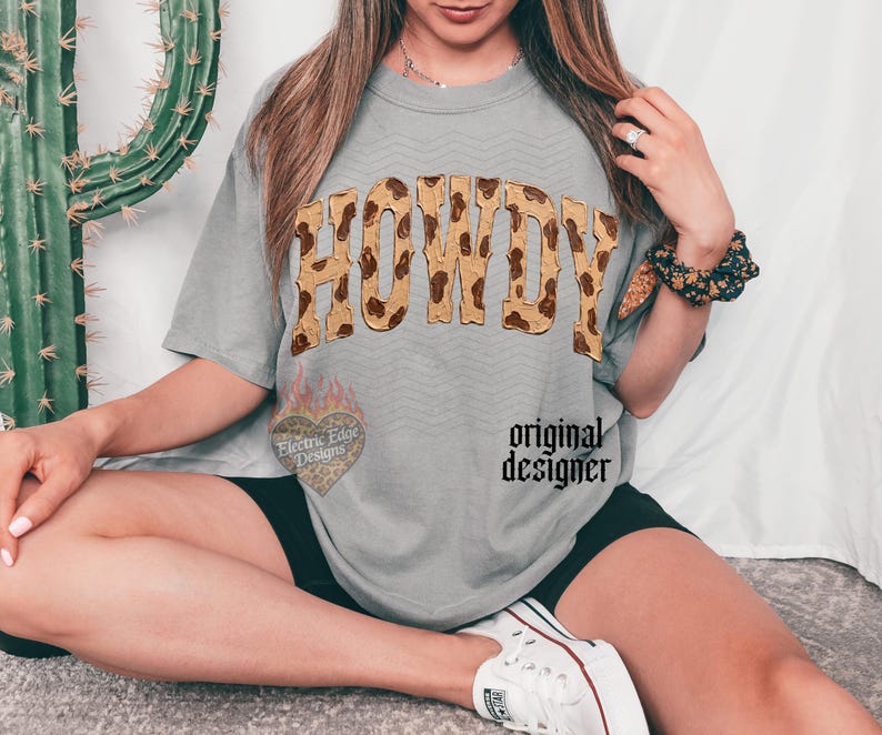 May include: A gray t-shirt with the word "HOWDY" in a brown and tan animal print design. The shirt also features a heart-shaped graphic with the text "Electric Edge Designs" and the words "original designer".