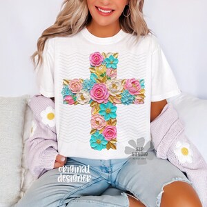 May include: White t-shirt featuring a floral cross design. The cross is adorned with pink, blue, and white flowers with gold accents. The shirt has a zig-zag pattern in the background and the words "original designer" are printed on the bottom left.