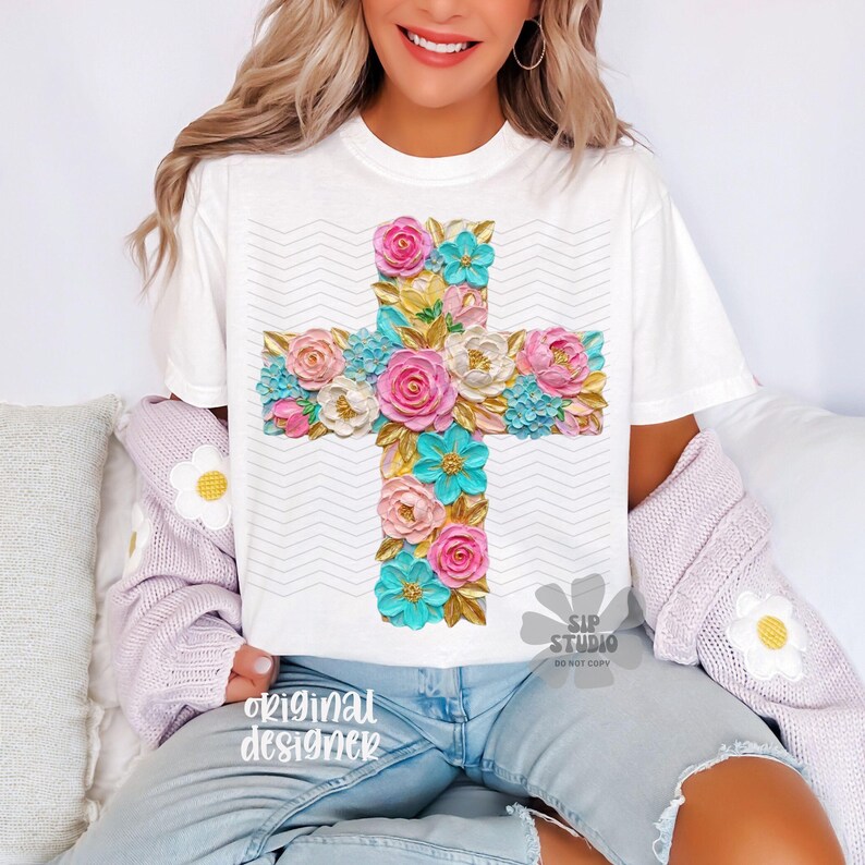 May include: White t-shirt featuring a floral cross design. The cross is adorned with pink, blue, and white flowers with gold accents. The shirt has a zig-zag pattern in the background and the words "original designer" are printed on the bottom left.