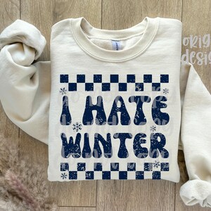 May include: Cream-colored sweatshirt with the phrase "I HATE WINTER" in navy blue, featuring a checkered pattern and snowflakes. The sweatshirt has long sleeves and a crew neck.