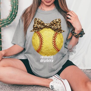 Op de afbeelding: Grijze t-shirt met een gele softbal, rode stiksels en een luipaardprint strik. De tekst "original designer" staat eronder. Het shirt is een casual, comfortabele stijl.