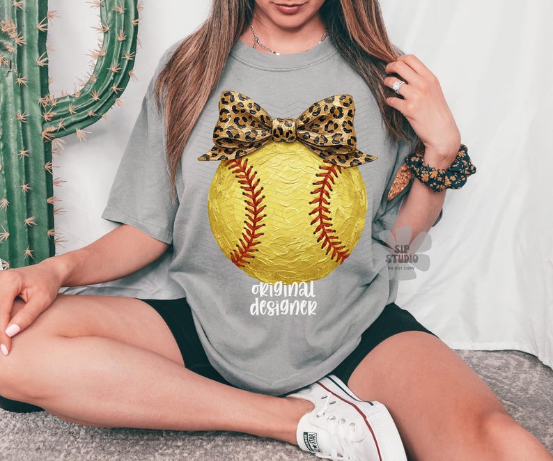 Op de afbeelding: Grijze t-shirt met een gele softbal, rode stiksels en een luipaardprint strik. De tekst "original designer" staat eronder. Het shirt is een casual, comfortabele stijl.