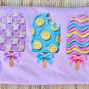 May include: A light purple t-shirt featuring a graphic of three popsicles. The left popsicle has a purple and white checkerboard pattern with a purple bow. The middle blue popsicle has yellow smiley faces and a blue bow. The right popsicle has wavy pink, blue, and yellow stripes with a pink bow.