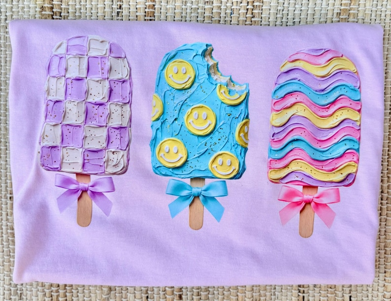 May include: A light purple t-shirt featuring a graphic of three popsicles. The left popsicle has a purple and white checkerboard pattern with a purple bow. The middle blue popsicle has yellow smiley faces and a blue bow. The right popsicle has wavy pink, blue, and yellow stripes with a pink bow.