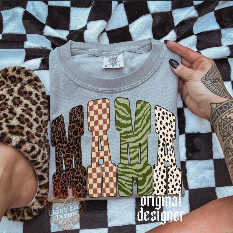 May include: A light gray crewneck t-shirt featuring the word MAMA in large block letters. Each letter has a distinct pattern: leopard print, brown and white checkerboard, olive green zebra stripes, and black and white dalmatian spots.
