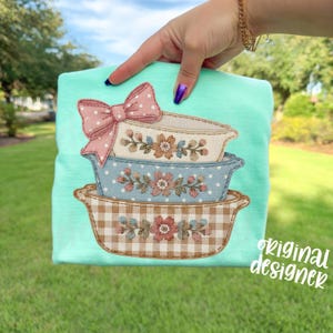 May include: A light turquoise t-shirt featuring an appliqu&eacute; design of three stacked, decorative baking dishes. The top dish has a pink bow with white polka dots. The dishes are decorated with floral and gingham patterns. The text "original designer" is visible.