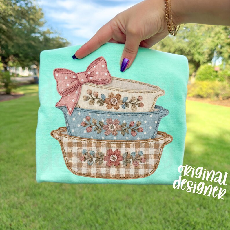 May include: A light turquoise t-shirt featuring an appliqu&eacute; design of three stacked, decorative baking dishes. The top dish has a pink bow with white polka dots. The dishes are decorated with floral and gingham patterns. The text "original designer" is visible.
