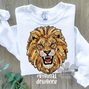 May include: White sweatshirt featuring a detailed, sequined lion's head design in shades of gold and brown. The lion has an open mouth, revealing teeth. The text "original designer" is printed at the bottom. The sleeves are folded.