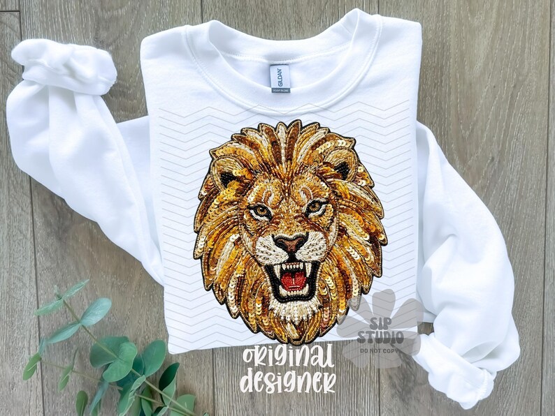 May include: White sweatshirt featuring a detailed, sequined lion's head design in shades of gold and brown. The lion has an open mouth, revealing teeth. The text "original designer" is printed at the bottom. The sleeves are folded.