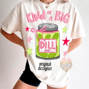 May include: Cream-colored t-shirt with a graphic of a pickle jar labeled "DILL PICKLES." The text "Kind of a BIG" is above the jar in pink, with star accents. The words "original designer" are below the jar.