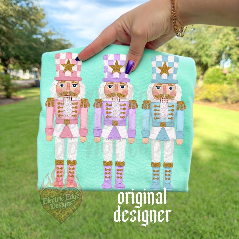 May include: A light mint green t-shirt featuring a graphic print of three nutcracker figures in pastel pink, lavender, and light blue. Each nutcracker wears a matching hat with a gold star, gold epaulets, and gold boots. The text reads Electric Edge Designs.
