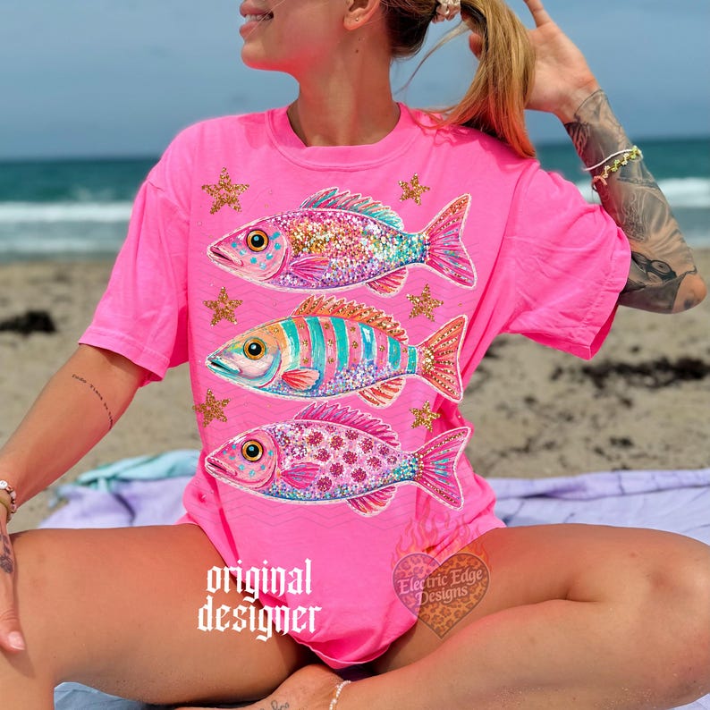 Puede incluir: Camiseta rosa brillante con tres dise&ntilde;os de peces coloridos con purpurina y estrellas. Los peces tienen patrones intrincados en tonos de rosa, azul y naranja. El texto "original designer" est&aacute; impreso en la camiseta.