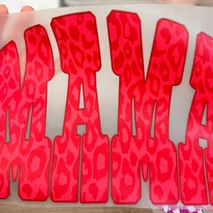 May include: A heat transfer decal featuring the word MAMA in large, bold, block letters. The design has a vibrant red and pink leopard print pattern throughout the text, set against a clear, thin, flexible adhesive backing material.