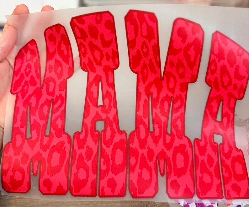 May include: A heat transfer decal featuring the word MAMA in large, bold, block letters. The design has a vibrant red and pink leopard print pattern throughout the text, set against a clear, thin, flexible adhesive backing material.