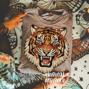 May include: A tan-colored t-shirt featuring a detailed tiger face design made with sequins and beads. The tiger has orange, brown, and black stripes, with a fierce expression. The text "original designer" is visible on the shirt.