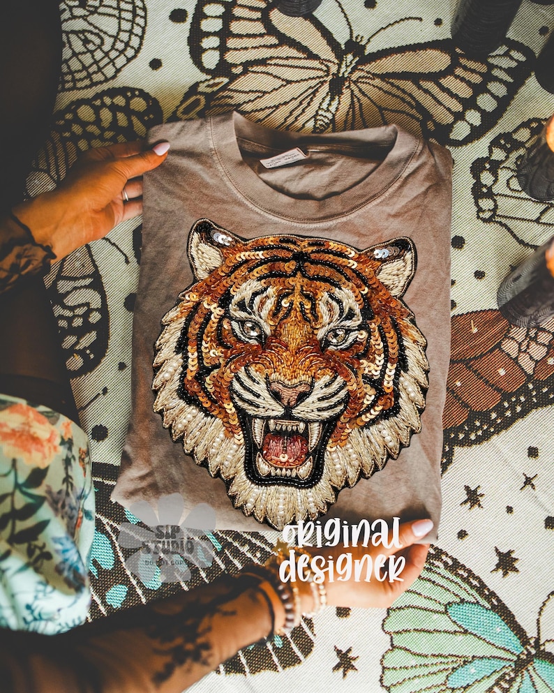 May include: A tan-colored t-shirt featuring a detailed tiger face design made with sequins and beads. The tiger has orange, brown, and black stripes, with a fierce expression. The text "original designer" is visible on the shirt.