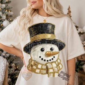 May include: A cream-colored short-sleeve t-shirt featuring a graphic of a snowman wearing a black top hat with gold glitter accents. The snowman has a textured orange carrot nose, black coal eyes and mouth, and a gold and white checkered scarf.