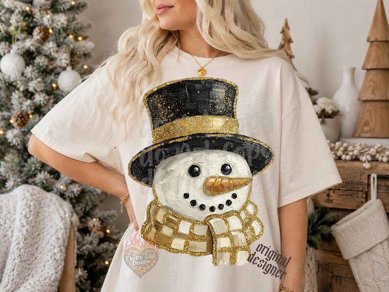 May include: A cream-colored short-sleeve t-shirt featuring a graphic of a snowman wearing a black top hat with gold glitter accents. The snowman has a textured orange carrot nose, black coal eyes and mouth, and a gold and white checkered scarf.