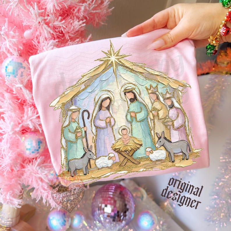 May include: A light pink t-shirt featuring a gold glitter-accented nativity scene illustration. The graphic depicts figures in pastel robes, a manger, a sheep, and a donkey under a star, with the text Electric Edge Designs printed below.