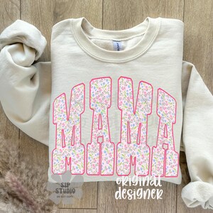 Op de afbeelding: Een beige sweatshirt met het woord "MAMA" in grote, roze omrande letters gevuld met een bloemenpatroon. De mouwen zijn opgerold en de achtergrond is een houten oppervlak.