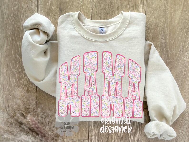 Op de afbeelding: Een beige sweatshirt met het woord "MAMA" in grote, roze omrande letters gevuld met een bloemenpatroon. De mouwen zijn opgerold en de achtergrond is een houten oppervlak.