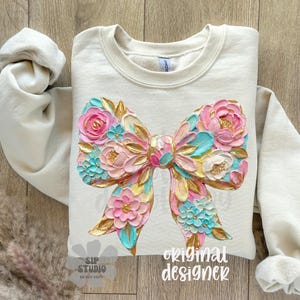May include: Cream-colored sweatshirt featuring a large, textured bow design. The bow is adorned with pink, blue, and gold floral elements. The words "original designer" are printed on the bottom right.