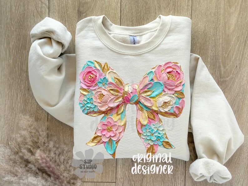 May include: Cream-colored sweatshirt featuring a large, textured bow design. The bow is adorned with pink, blue, and gold floral elements. The words "original designer" are printed on the bottom right.