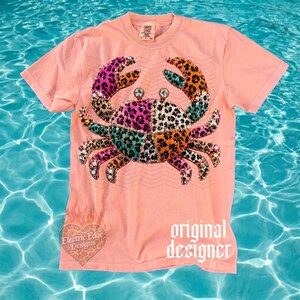 May include: A coral-colored t-shirt featuring a crab design with a patchwork pattern in pink, teal, orange, and leopard print. The crab has rhinestone accents and is centered on the shirt. The words "original designer" are printed on the bottom right.