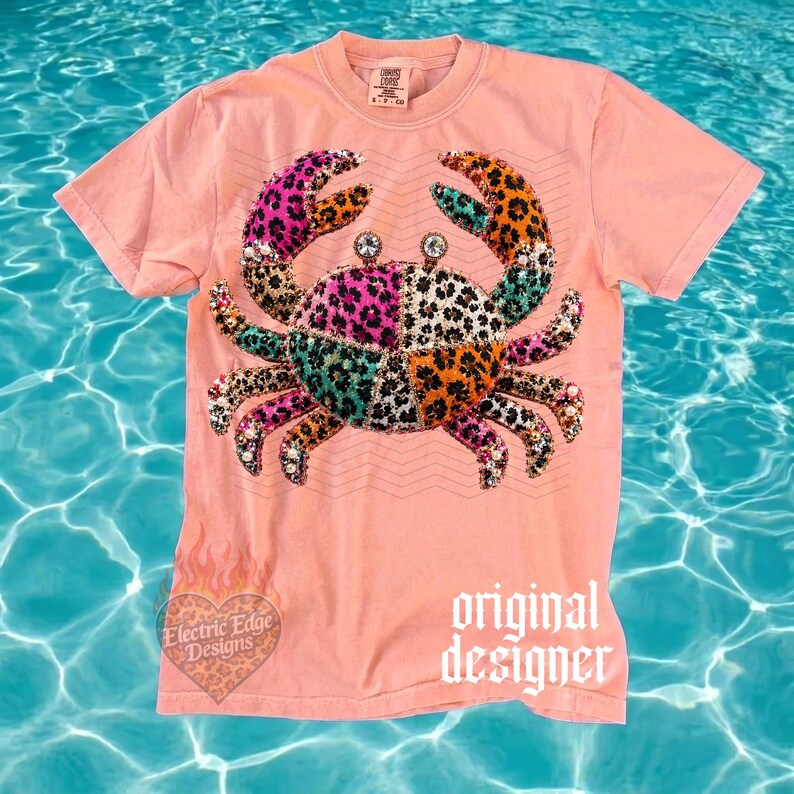 May include: A coral-colored t-shirt featuring a crab design with a patchwork pattern in pink, teal, orange, and leopard print. The crab has rhinestone accents and is centered on the shirt. The words "original designer" are printed on the bottom right.