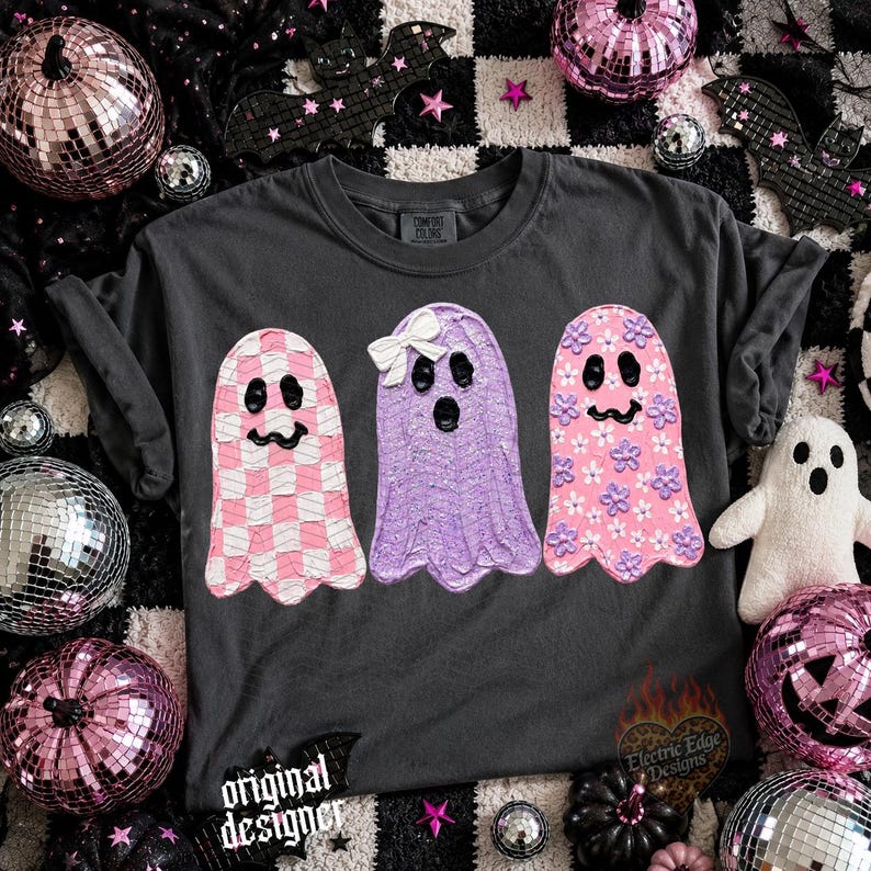 May include: A charcoal gray t-shirt featuring three ghost illustrations. The left ghost has a pink and white checkered pattern, the middle ghost is solid purple with a white bow, and the right ghost is pink with small purple and white flower accents.