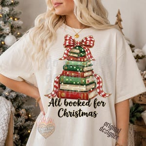 May include: An off-white short-sleeve t-shirt featuring a graphic of a Christmas tree made of stacked books topped with a red and white plaid bow. The text All booked for Christmas is printed in black below the book stack illustration.