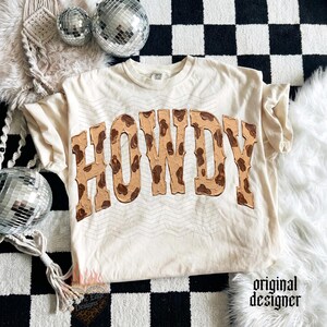 May include: Cream-colored t-shirt with the word "HOWDY" in a brown, textured font. The shirt has a relaxed fit and short sleeves. The text has a cowhide pattern. The shirt is on a black and white checkered surface.