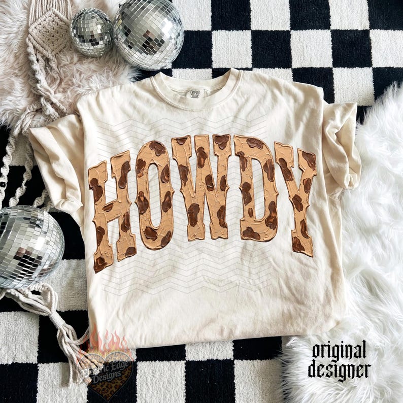 May include: Cream-colored t-shirt with the word "HOWDY" in a brown, textured font. The shirt has a relaxed fit and short sleeves. The text has a cowhide pattern. The shirt is on a black and white checkered surface.