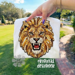 May include: White t-shirt featuring a detailed, sequined lion's head design in shades of gold, brown, and black. The lion has an open mouth with visible teeth. The text "ORIGINAL DESIGNER" is printed on the shirt.
