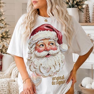 Puede incluir: Una camiseta blanca de manga corta con un gr&aacute;fico texturizado de estilo empaste que muestra a Santa Claus con un gorro rojo. El dise&ntilde;o incluye una barba y un bigote blancos y gruesos con pinceladas visibles. El texto "original designer" est&aacute; impreso en dorado.