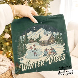 May include: A dark green sweatshirt featuring a graphic print of a snowy mountain landscape with log cabins, people ice skating, and a campfire. The text "WINTER VIBES" is printed in a cream-colored script font at the bottom of the design.