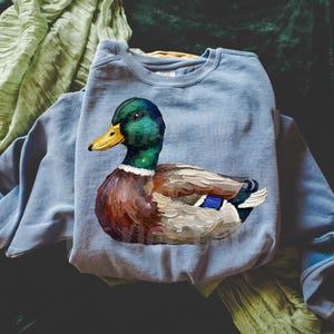 May include: A light blue sweatshirt featuring a detailed painting of a mallard duck. The duck has a green head, yellow beak, and brown and white body. The sweatshirt is made of a soft material.