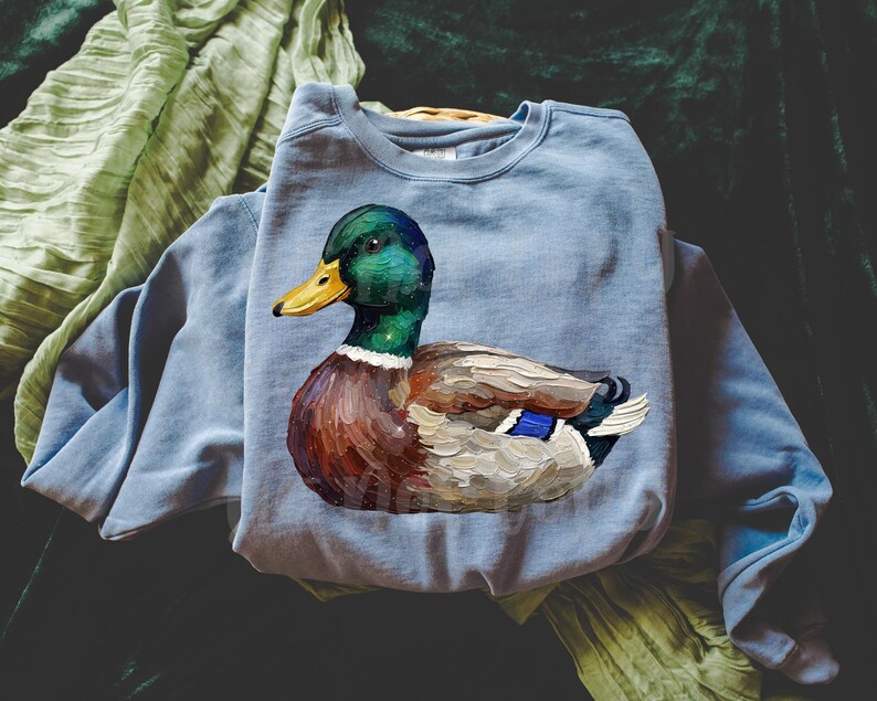 May include: A light blue sweatshirt featuring a detailed painting of a mallard duck. The duck has a green head, yellow beak, and brown and white body. The sweatshirt is made of a soft material.