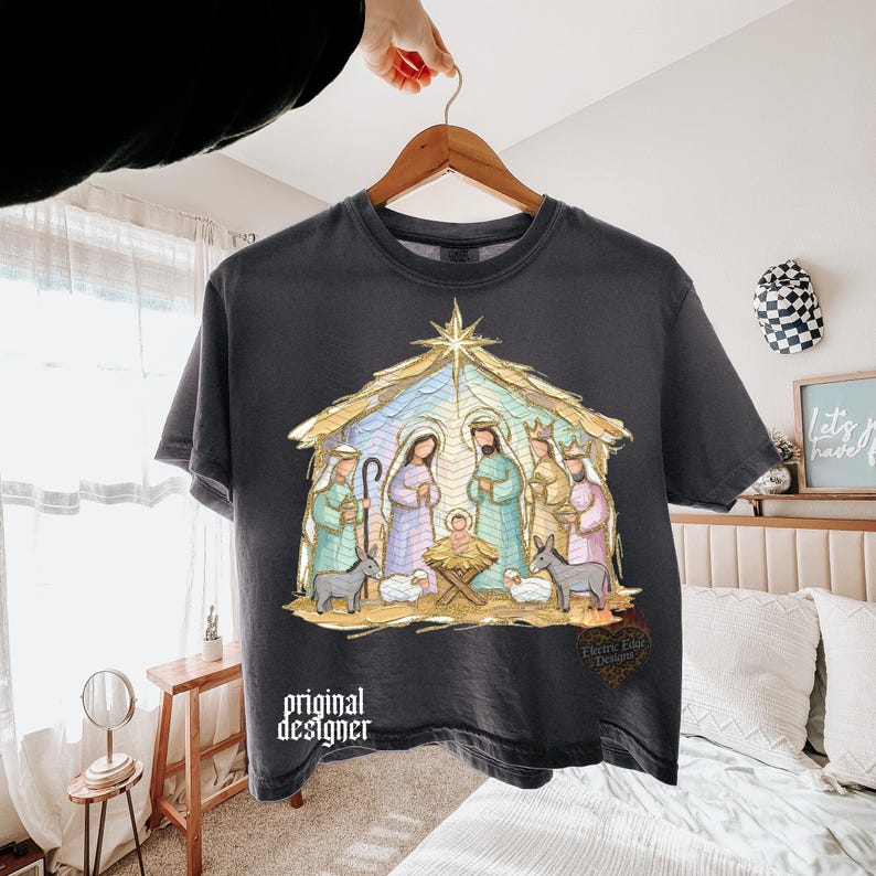 May include: A charcoal gray short-sleeve t-shirt featuring a colorful graphic of a nativity scene with a star above, figures in pastel robes, and animals. The text "original designer" is printed in white on the lower left side of the shirt.