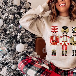 Peut inclure: Un sweat-shirt beige avec trois illustrations color&eacute;es de casse-noisettes. Les casse-noisettes portent des uniformes rouges, blancs et dor&eacute;s avec des bottes noires. Un sapin de No&euml;l avec des boules blanches est en arri&egrave;re-plan.