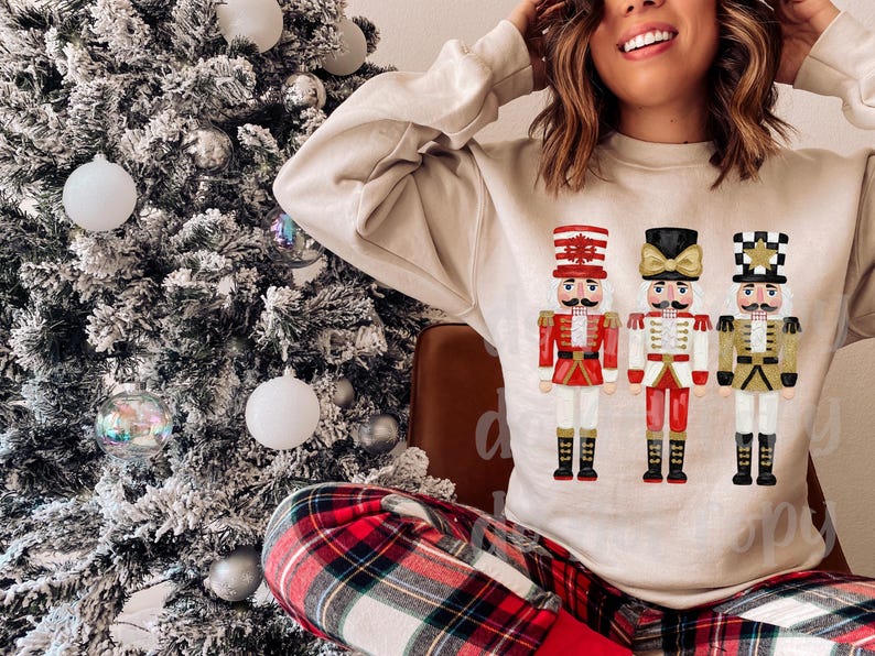 Peut inclure: Un sweat-shirt beige avec trois illustrations color&eacute;es de casse-noisettes. Les casse-noisettes portent des uniformes rouges, blancs et dor&eacute;s avec des bottes noires. Un sapin de No&euml;l avec des boules blanches est en arri&egrave;re-plan.