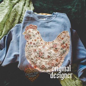 May include: A light blue sweatshirt with a floral chicken applique. The chicken design features a patchwork style with a variety of floral patterns in shades of pink, cream, and green. The text "original designer" is visible.