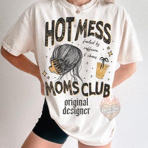 May include: Cream-colored t-shirt with the text "HOT MESS MOMS CLUB" in black and yellow. The shirt features a graphic of a woman's hair, a coffee cup, and the words "fueled by caffeine & chaos".