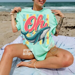 May include: Mint green t-shirt with a large graphic of a blue whale and the word "Oh" in red script. The design includes yellow stars and vertical stripes. The words "original designer" are printed on the shirt.